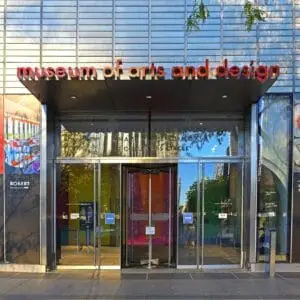 Entrance to the Museum of Arts and Design with large glass doors and a red sign above. Posters for Fashion in NYC - Grade 12 (March) are displayed on both sides, with some greenery reflected in the glass. Educational Tours | Travel for Credit | Student Travel Programs | edutravelforcredit.com | Phone: 416-923-4638 | Toll Free: 1-877-523-4638