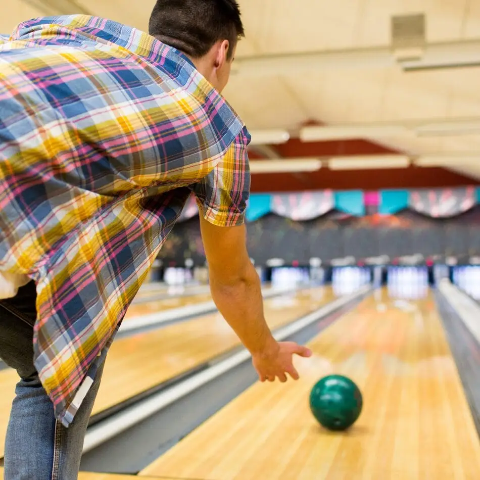 A person wearing a plaid shirt is seen from behind, about to release a green bowling ball down a wooden bowling lane in a brightly lit bowling alley, reminiscent of the teamwork and focus seen in CPS: Ski BC - Grade 12 (March) events. Educational Tours | Travel for Credit | Student Travel Programs | edutravelforcredit.com | Phone: 416-923-4638 | Toll Free: 1-877-523-4638