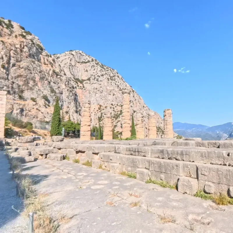 Archaeological site of Delphi in Greece, featuring ancient stone ruins and columns set against a dramatic mountain landscape under a clear blue sky.
