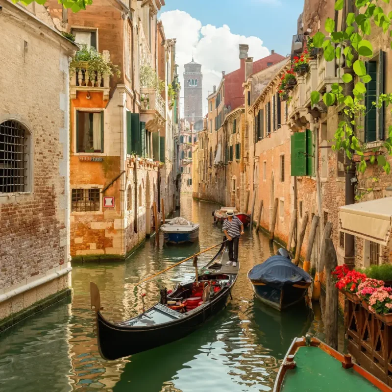Gondola travelling along a narrow canal in Venice, surrounded by historic buildings and colourful flowers.